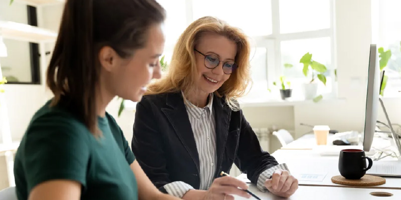 Two women sitting at a desk, using a pen and paper to jot down notes and ideas.